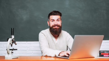 man sitting at a table typing on a laptop in front of a chalkboard