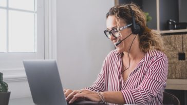 woman sitting at a work table typing on a laptop and talking on headphones