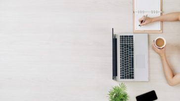 bird view of laptop, phone, plant, coffee and someone writing in a notebook