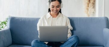 smiley woman with headphones sitting on a couch and working on a laptop